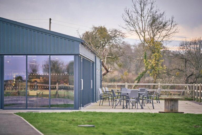A farm style building housing a cafe, with tables and chairs outside on the terrace and trees in the distance
